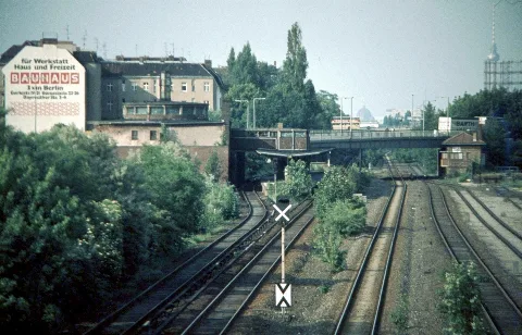 To the left, in front of the Feuerbach Bridge, lies the Feuerbachstraße S-Bahn station on the Wannsee Line, which has been out of service since 1980. To the right of this are the tracks of the former Stammbahn, and further right is the Steglitz freight yard, situated between Albrechtstraße and the Feuerbach Bridge, along with the "Sob" signal box. Also visible are the Schöneberg gasometer and, on the horizon in East Berlin, the Berlin Cathedral and the TV Tower at Alexanderplatz.