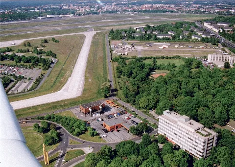 At the bottom left is the access road junction. In the center of the image lies the public gas station, and above it, within the secure area, is the grit storage facility for the airport’s winter maintenance service. To the right of that stands the administrative building of Berlin Airport GmbH. In the center-left of the image is the public parking lot P4, extending to the right is taxiway PE, and to the right of that, the internal connecting road to Tegel North. In the public area on the right, one sees the Novotel, and above it, the site used for the German-French fair. Further up again is the ADAC driver training area, followed by Cité Pasteur, a residential complex for the French occupying forces. To the right of this runs Kurt-Schumacher-Damm, with Quartier Napoléon, headquarters of the French armed forces in Berlin, located immediately to its right.