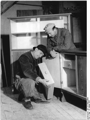 For documentary purposes the German Federal Archive often retained the original image captions, which may be erroneous, biased, obsolete or politically extreme. Rhön, Möbelfabrik, Endkontrolle Zentralbild Bez.Red.Erfurt Br-Qu 20.1.1956 Bessere Möbel durch Wettbewerb. Ein verbessertes Sortiment an Schlafzimmer- und Zusatzmöbeln wird im kommenden Jahr die Tischlereibetriebe der Genossenschaft des Kunst- und Holzhandwerks, Kaltennordheim in Rhön, Kreis Bad Salzungen, verlassen. Die Genossenschaft haben sich etwa 340 (drei vier null) Betriebe angeschlossen. Entscheidend für das verbesserte Warensortiment war ein Wettbewerb, den die Mitglieder der Genossenschaft bereits seit 1954 erfolgreich durchführen. Schon jetzt tragen die Möbel der Genossenschaftsbetriebe das Gütezeichen. Mit Aufnahme der neuen Modelle liefert die Genossenschaft rund 80 (acht null) % ihrer Gesamtproduktion nach Sonderklasse und Güteklasse 1. Die Holzhandwerker aus der Rhön haben alle artverwandten Betriebe des Bezirkes Suhl im November 1955 aufgefordert, ihrem Beispiel zu folgen. Dieser Aufruf ist von den Holzhandwerksgenossenschaften des Bezirkes Suhl aufgegriffen worden. Die Handwerkskammer des Bezirkes Suhl will einen Wettbewerb organisieren und hat Prämien in Höhe von DM 5 000,-- (fünf null null null) zugesagt. In einer Leistungsschau in Bad Salzungen werden im April dieses Jahres die Genossenschaftsmitglieder von Kaltennordheim für den Wettbewerb bereits ihre Produktionsmuster für 1957 ausstellen. UBz: In der Werkstatt des Siegerbetriebes vom Vorjahr. Der Tischlermeister Helmut Gerlach erhielt im Wettbewerb 1955 den 1. Preis auf ein Wohnzimmer, welches mit dem Gütezeichen S ausgezeichent wurde. Die gesamte Produktion des Betriebes liegt in Güteklasse S und Güteklasse 1. Qualitätsprüfer der Genossenschaft. Tischlermeister Kurt Bruckner (links) bei der Gütekontrolle beim Tischlermeister Helmut Gerlach (rechts). Dieses Wohnzimmer erhielt den 1. Preis im Wettbewerb 1955 und als erstes in der Genoss