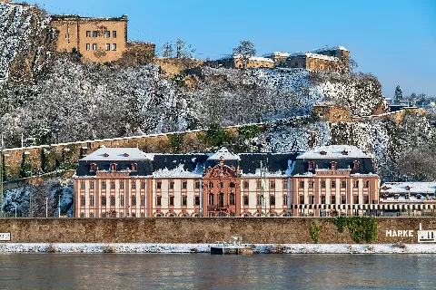 Dikasterialgebäude und Festung Ehrenbreitstein im Winter I. Das Dikasterialgebäude: Der Kurstaat Trier als Landesherr Die winterliche Aufnahme dokumentiert das architektonische Spannungsfeld am rechten Rheinufer zwischen barocker Verwaltungsarchitektur und monumentaler Festungsbaukunst. Im unteren Bildzentrum dominiert das schlossartige Dikasterialgebäude das Rheinufer: Ein zentraler Regierungsbau: Das Gebäude wurde zwischen 1739 und 1749 im Auftrag des Trierer Fürsterzbischofs und Kurfürsten Franz Georg von Schönborn errichtet. Nachdem die Verwaltung des Kurstaates Trier für über ein halbes Jahrhundert nach Ehrenbreitstein verlegt worden war, bündelte dieser monumentale Bau die kurfürstliche Regierung und das Obergericht unter einem Dach. Die Handschrift zweier Baumeister: Die architektonischen Entwürfe für den langgestreckten, dreigeschossigen Trakt stammen von dem berühmten Barockbaumeister Balthasar Neumann. Die praktische Bauleitung vor Ort übernahm ab 1739 der kurtrierische Hofarchitekt Johannes Seiz, der das monumentale Bauwerk mit seinen 25 Achsen und dem prachtvoll verzierten Mittelrisaliten vollendete. Ein überlebendes Denkmal: Das Dikasterialgebäude stand ursprünglich in direkter Nachbarschaft zur Philippsburg, der Residenz der Kurfürsten. Während das eigentliche Schloss im Jahr 1801 von französischen Truppen gesprengt wurde, blieb der Verwaltungsbau als wichtiges Zeugnis kurtrierischer Landesherrschaft erhalten. Die moderne Zäsur durch die Eisenbahn: Unmittelbar vor der historischen Ufermauer des Dikasterialgebäudes verlaufen die Gleisanlagen der rechten Rheinstrecke. Direkt rechts an das Gebäude anschließend erstreckt sich der Bahnhof Koblenz-Ehrenbreitstein, dessen Bahnsteigdach und Gleiskörper im Vordergrund sichtbar sind. Diese verkehrstechnische Infrastruktur des 19. Jahrhunderts trennt seither die barocke Schaufassade vom direkten Zugang zum Flussufer. II. Die Festung Ehrenbreitstein: Das preußische Bollwerk am Rhein Auf dem steilen Felsrücken ober