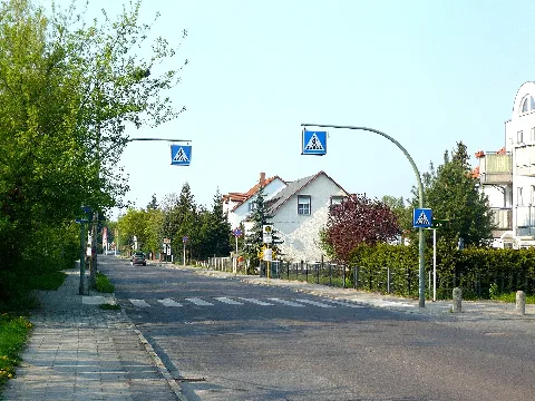 Berlin-Kaulsdorf in der Kaulsdorfer Straße mit gezoomtem Blick nördlich. Der Standort ist an der Ecke zur Bismarcksfelder Straße. Voraus ist rechts an der Grundstücksecke das Ortsschild für Kaulsdorf zu sehen, aber auch der Bereich am Standort gehört zu Kausdorf dazu. Wenig hinter dem Betrachter verläuft der „Eichwaldgraben“ (vermutlich auch abseits der Straße verrohrt unter der Erde) schräg über die Straße hinweg zur Wuhle, dies ist die Grenzlinie zwischen Köpenick und Kaulsdorf. Dies ist zwischen Nr. 93 und 95 auf der rechten und Nr. 98 und 94 (Supermarkt) auf der linken Seite. Vor dem Fußgängerüberweg geht rechts die Feuersteiner Straße ab. Das angeschnitten zu sehende Gebäude rechts an der Bildkante ist die Kaulsdorfer Straße 87. Links, wo auch die gepflasterte Zufahrt hin führt, liegt nur als üppiger Grünwuchs erkennbar das Gelände vom ehemaligen „Zwangsarbeiterlager“ / „Lager Kaulsdorfer Straße 90“. Von diesem ist nur eine Baracke verblieben, welche heutzutage als Privathaus dient. Der Rest des Geländes ist als Eigenheimsiedlung mit der Straße „Am Wuhlebogen“ als Zubringer längs durch den Streifen, von der Kaulsdorfer Straße abgekapselt bebaut worden. Die Grundstückszäune stehen heutzutage direkt an der Befestigung des Fußweges, welche sich laut Google Streetview Bildern von Aprill 2022 dort noch so erhalten hat. Auch die Pflasterung der Einfahrt ist danach noch erhalten geblieben, es gibt dort aber kein Tor im Zaun. Dort liegt heutzutage das Grundstück Am Wuhlebogen 10. Ebenso hat sich bis mindestens zu diesem Zeitpunkt die Straßenlaterne links hinter dem Überweg erhalten. Diese stammt noch aus DDR Zeiten und trägt am Doppelausleger auf ihrem Betonmast zwei „BG 0“ Leuchten vom VEB Leuchtenbau Leipzig. Rechts hinter dem Überweg liegt die Bushaltestelle „Feuersteiner Straße“. Nach links herüber verläuft hinter dem Siedlungsstreifen nur ca. 105m entfernt die Wuhle, welche auf Höhe der Bismarcksfelder Straße für Fußgänger und Radfahrer per Bismarcksfelder Brücke