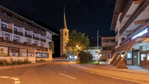 Kirchdorf in Tirol by night. As streets you can see the village square and the village street. On the right you can see the Raiffeisenbank Kirchdorf Tirol, on the left behind it the Spar and in the center of the picture the parish church St. Stephan. On the left you can see a kind of inn with a youth club.