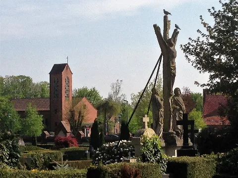 Im Vordergrund ist die Kreuzigungsgruppe auf dem Friedhof der katholischen Pfarrgemeinde St. Gertrud in Lohne (Landkreis Vechta, Niedersachsen) zu sehen. Im Hintergrund steht die evangelische Kirche St. Michael.