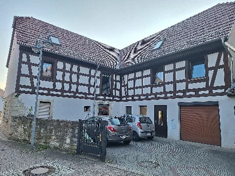Courtyard view of the residential building at Mittelstraße 2 in Bad Klosterlausnitz, Saale-Holzland district, Thuringia, Germany. The two-storey, L-shaped building has a rendered masonry ground floor and an upper floor with exposed timber framing and white rendered infill panels. Its two wings have red-tiled gable roofs and enclose a paved courtyard opening towards Mittelstraße. A low natural-stone wall and a metal gate stand along the street, while the ground floor of the right-hand wing includes a garage.