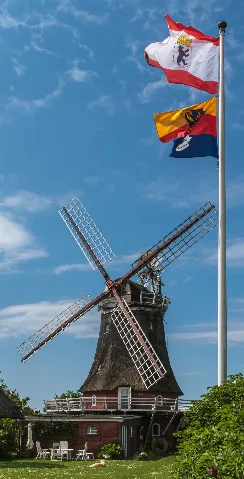 This is a photograph of a cultural monument.It is on the list of cultural monuments of Oldsum-Toftum, no. 3512. Designation: The Oldsum windmill with flags in the wind Construction time: 1901 Description: Galleried Dutch windmill, rebuilt to a residential building Place: Quarter Toftum, Local municipality Oldsum, Collective municipality Föhr-Amrum, District Nordfriesland, Schleswig-Holstein, Germany Location: Eemelkeswai House number: 190