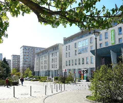 Berlin-Marzahn, an der Sella-Hasse-Straße, nahe der Mehrower Allee (im Rücken des Betrachters). Der Standort ist an eine Schleife seitlich der Straße, welche als Taxistand dient, der Blick geht süd-westlich zum „Plaza Marzahn“. Links geht der Weg der „Ringkolonnaden“ hinein, dessen verlängerter Verlauf rechts auch durch das Plaza führt. Links vom Standort, außerhalb des Bildbereiches liegt eine Grünfläche, welche eher einer Brache gleicht, mit mehreren hindurchlaufenden Trampelpfaden. Hinter dieser liegt dann das Ärztehaus (ehemals DDR Poliklinik. Auf dieser Grünfläche wurde dann rund 10 Jahre später das Hochhaus Mehrower Allee 20a, der „Ella Wohnturm“ errichtet.)