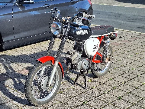 A Simson S51 moped with a black fuel tank, white side covers, and red mudguards parked in the parking lot of the McDonald's restaurant in Petersberg (Aubitz), Saale-Holzland district, Thuringia, Germany. The motorcycle is standing on its centre stand and is shown from the front left at an angle. A parked passenger car and the paved parking area are visible in the background.