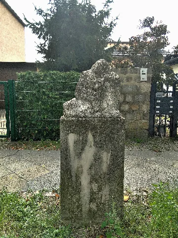 Sitzende Figur in der Wernerstraße in Berlin-Kaulsdorf. Diese steht auf dem grünen Straßenrandstreifen gegenüber der abgehenden Ernst-Haeckel-Straße zwischen den Einfahrten der Grundstücke Nr. 17 und 15, welche im Hintergrund zu sehen sind.