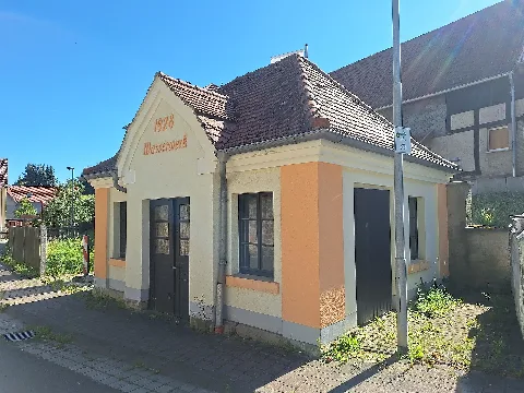 Oberndorf waterworks building on Klosterlausnitzer Straße in Oberndorf, a district of the municipality of Kraftsdorf, Greiz district, Thuringia, Germany. The photograph shows the small single-storey rendered utility building with a tiled hipped roof, a central gable above the entrance, and dark-painted doors and windows. The raised inscription “1928 Wasserwerk” (“1928 Waterworks”) on the gable indicates the period in which the building was constructed.